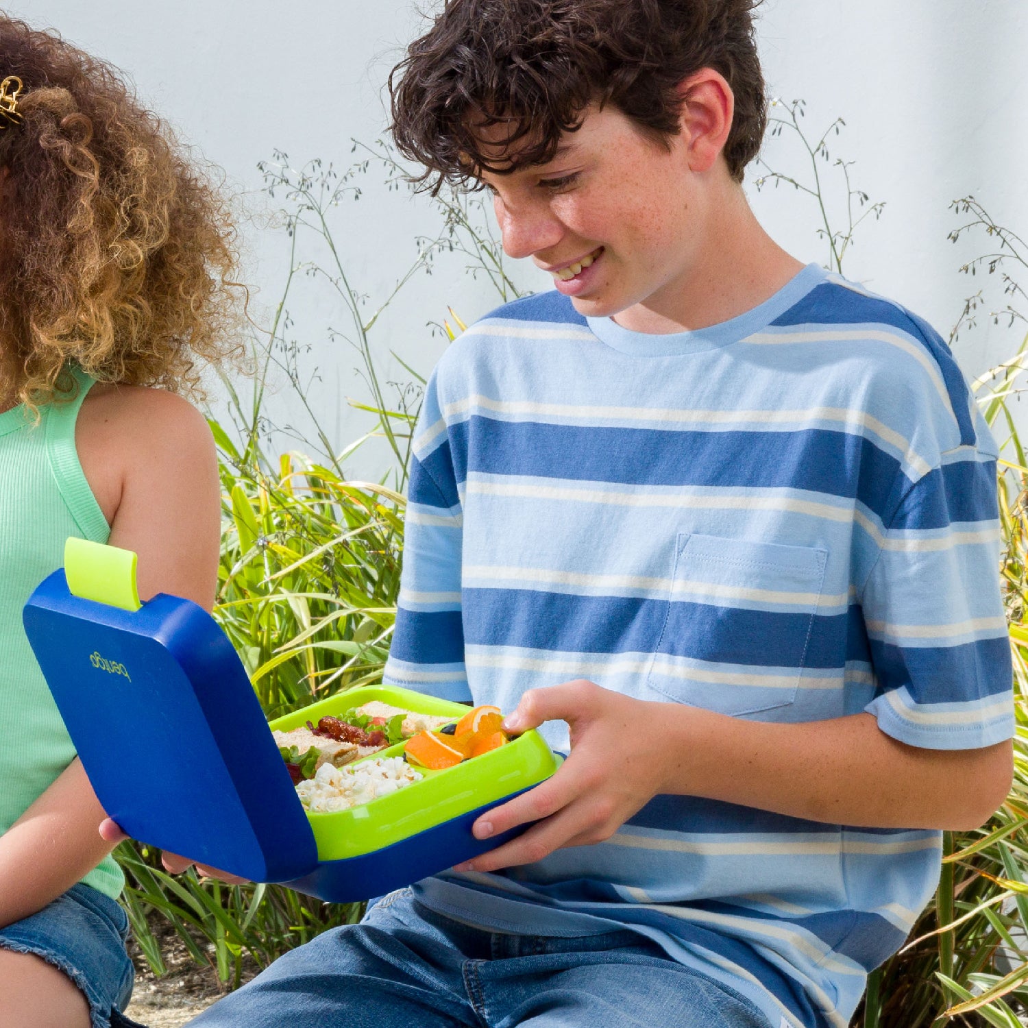 Two children sitting outdoors with a blue and green lunch box containing food.