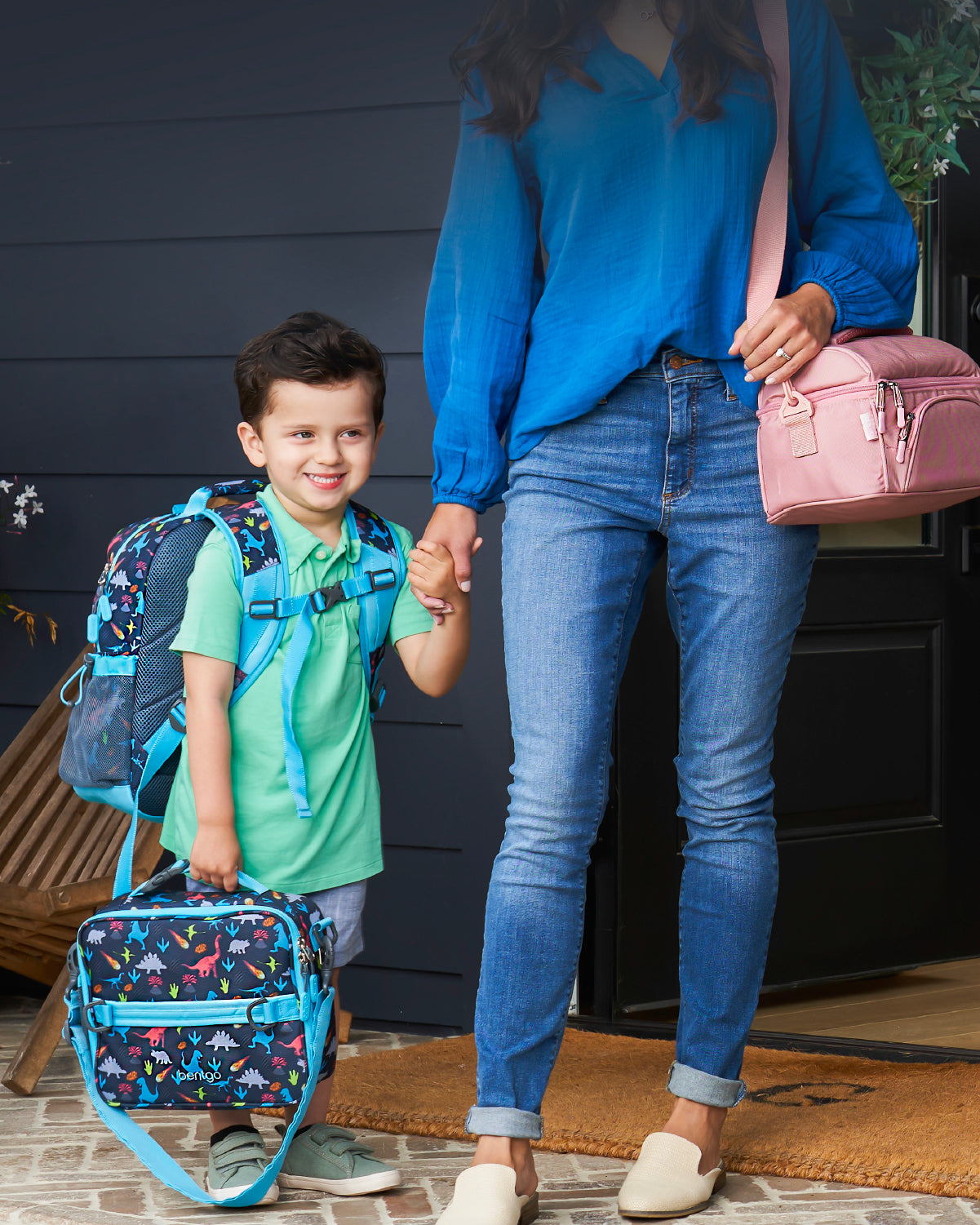 Child with a backpack and colorful bag, holding hands with an adult, standing on a porch.