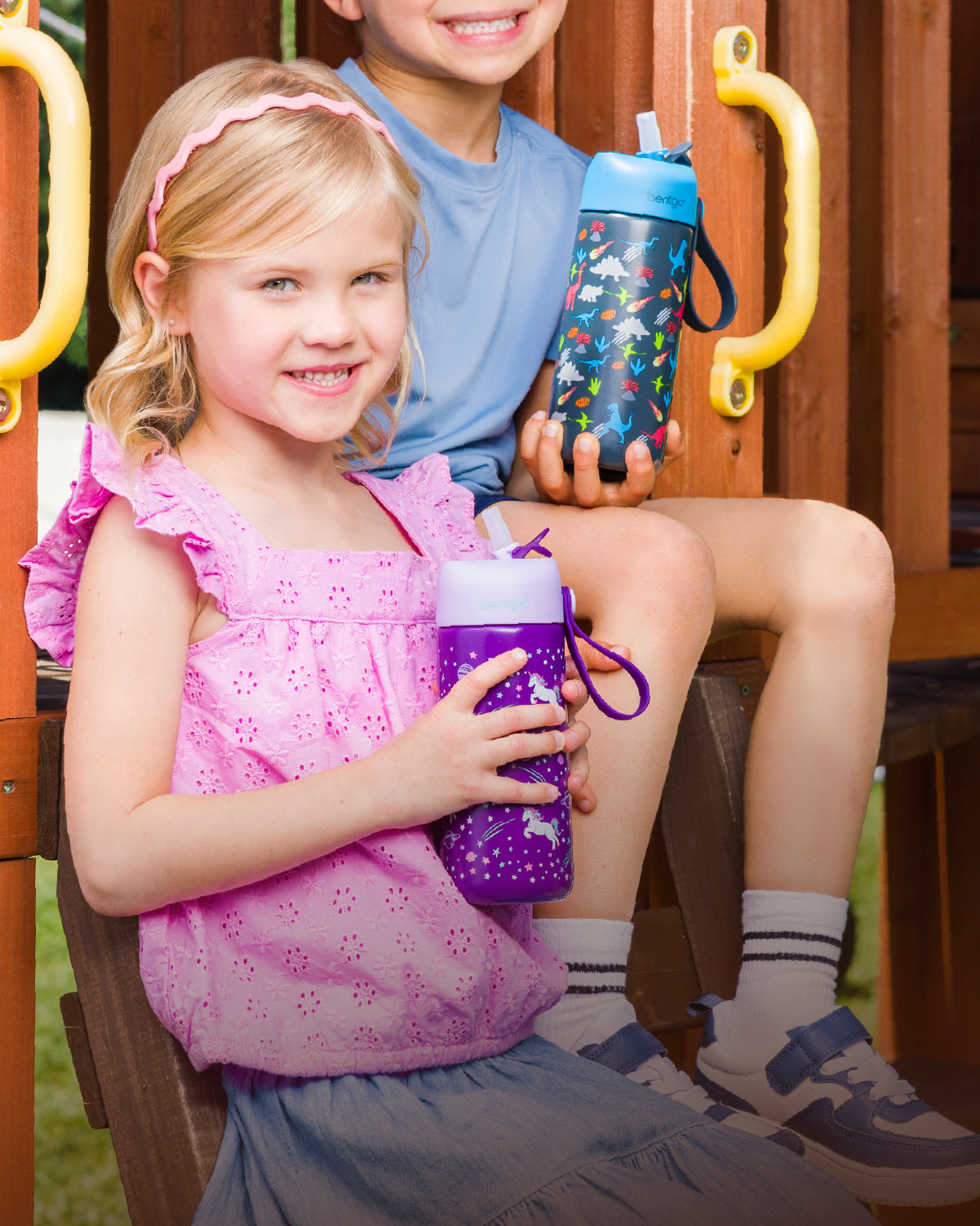 Two children sitting on a wooden slide holding colorful water bottles.